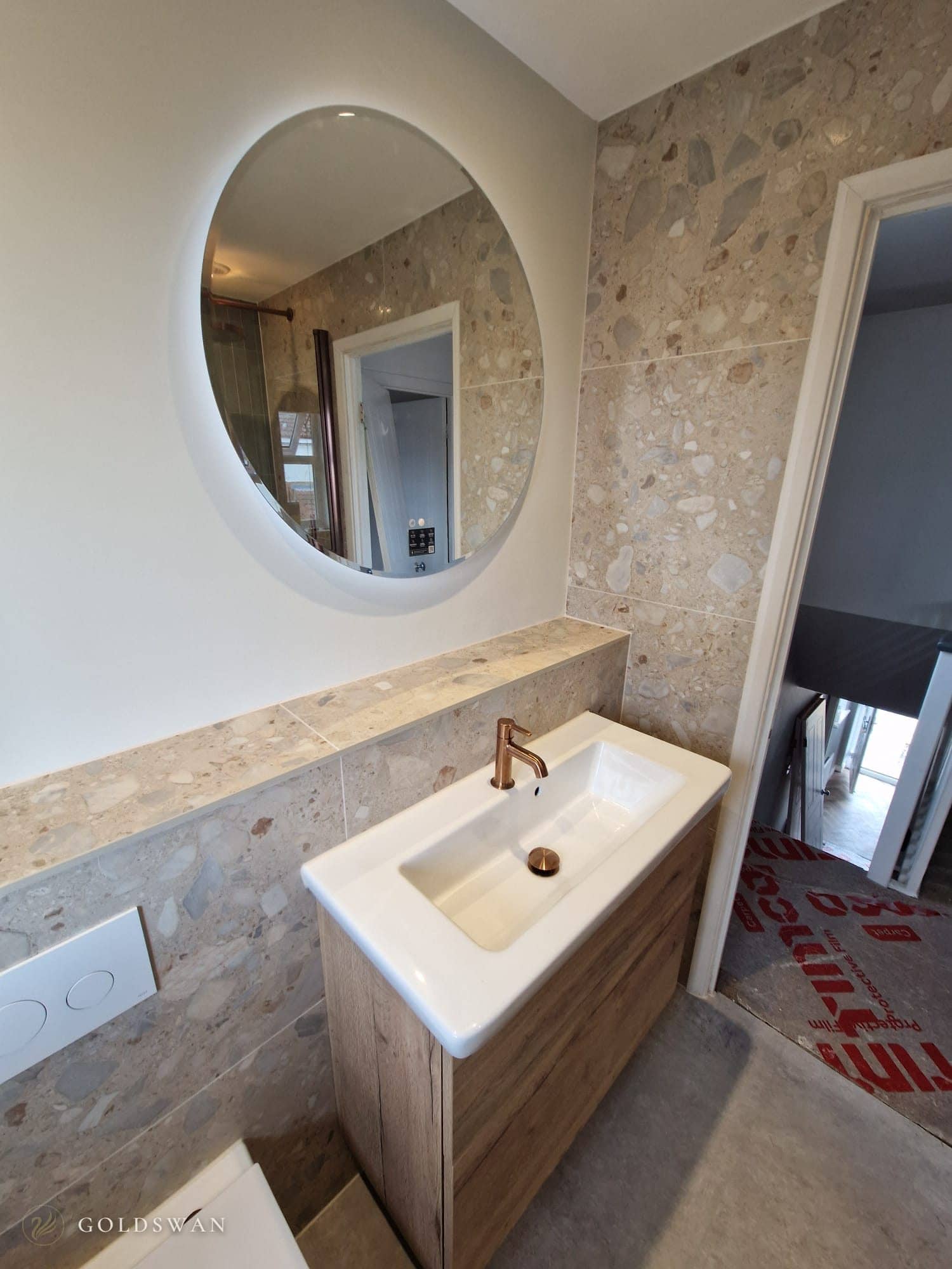 Round backlit mirror above a wall hung oak effect vanity with a white basin and brushed copper tap, with a terrazzo effect tiled ledge and wall