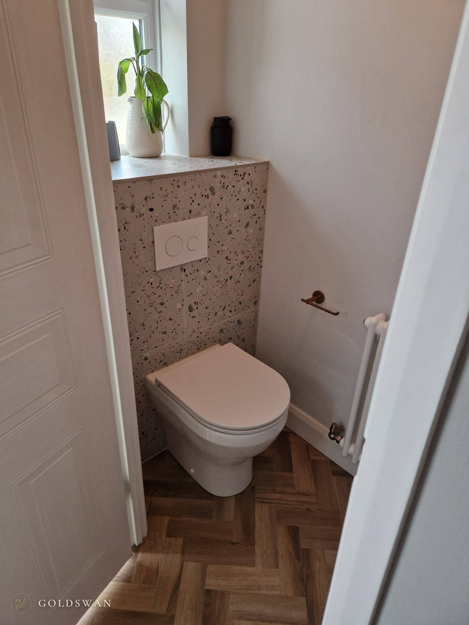 Downstairs WC with a back to wall toilet against multicoloured terrazzo effect tiles, a white flush plate, a brushed copper roll holder, a white column radiator and herringbone wood effect flooring