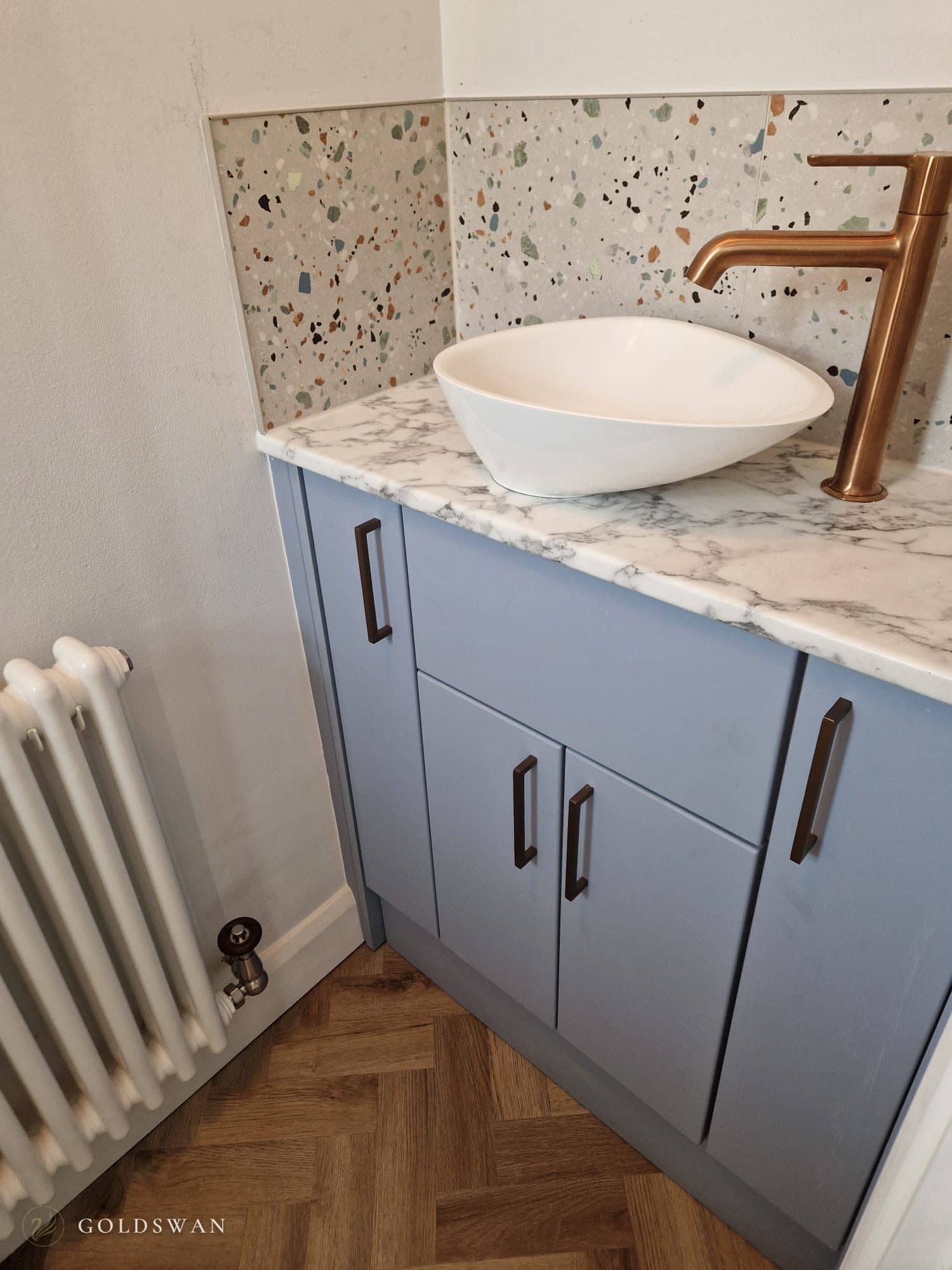 Pale blue cloakroom vanity with a marble effect top, a white countertop basin and a brushed copper tap, against multicoloured terrazzo effect tiles beside a white column radiator