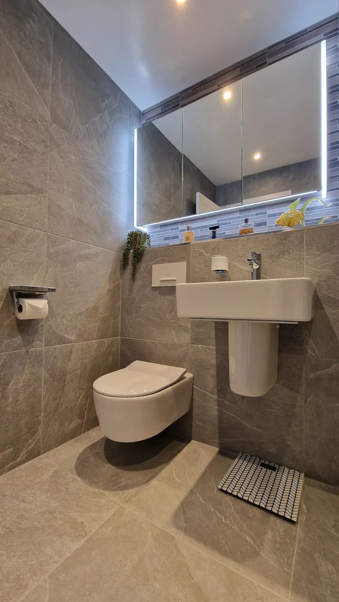 Corner of a grey stone effect room with a wall hung toilet, a white semi pedestal basin hung on the wall with a chrome tap, a white flush plate, and a mirror cabinet with lit edges above a tiled ledge