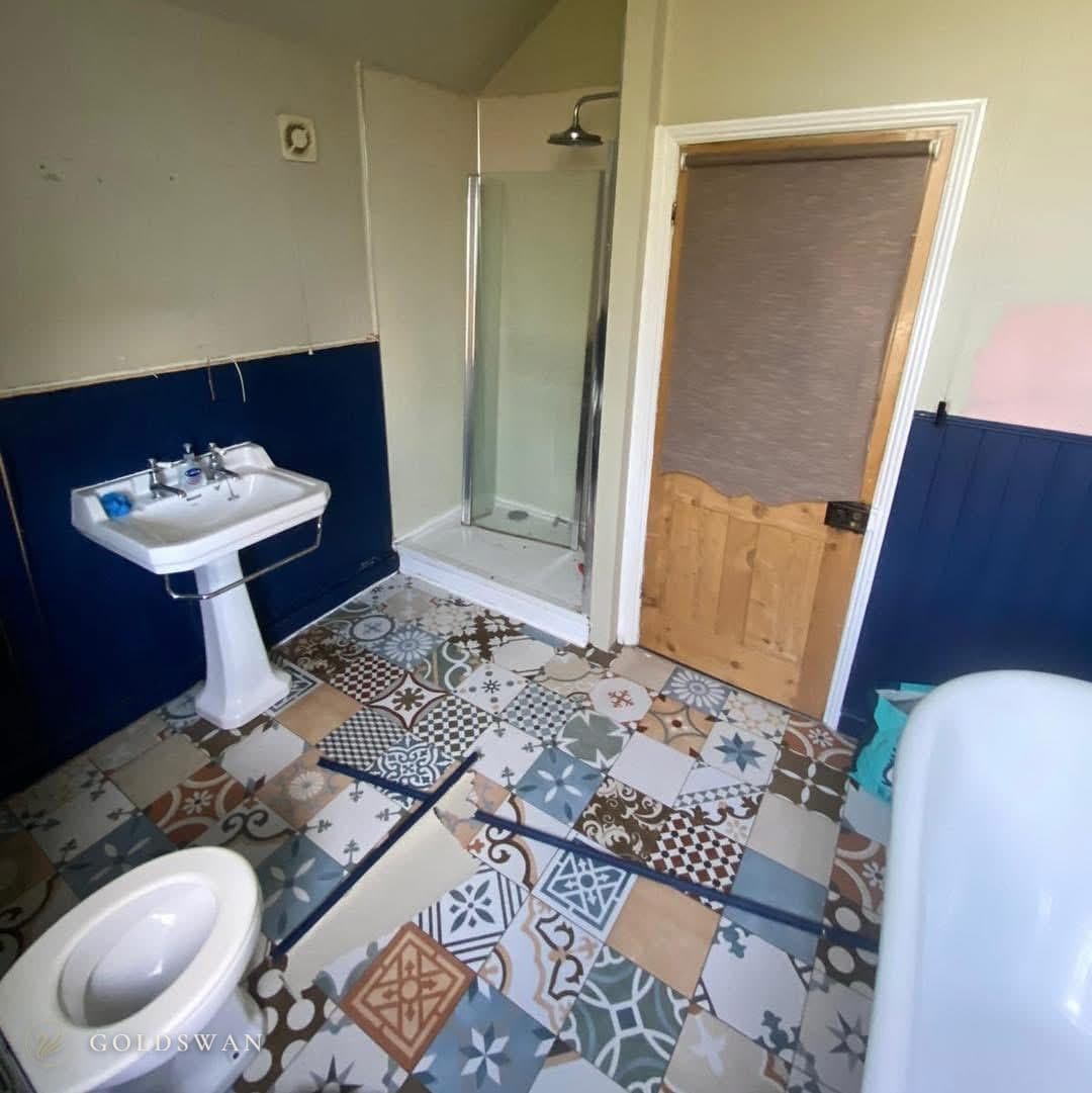 The bathroom before: navy tongue and groove panelling to dado height, a pedestal basin, a corner shower cubicle beside a wooden door and a patchwork of patterned floor tiles