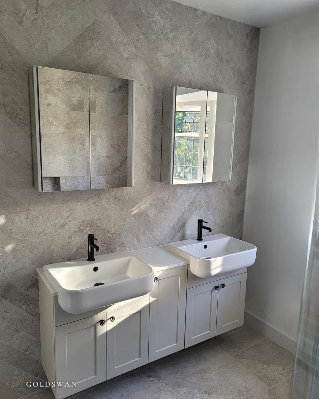 Two semi-recessed basins with matt black taps on pale shaker style units, each under its own mirror cabinet, against grey stone effect tile laid in herringbone