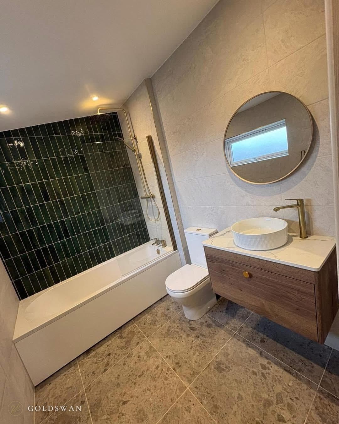 Bath with a brushed brass hinged screen and an exposed brushed brass shower riser with a rain head and handset, against deep green gloss tiles laid in narrow vertical stacks, beside a close coupled toilet and a wall hung wood effect vanity with a round countertop basin, a tall brass mixer and a round brass framed mirror