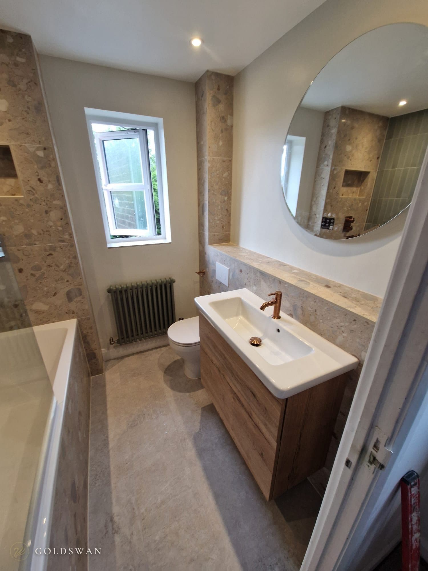 Family bathroom with a bath and glass screen, terrazzo effect stone feature walls, a sage green column radiator under the window, a wall hung oak effect vanity with a brushed copper tap and a round backlit mirror