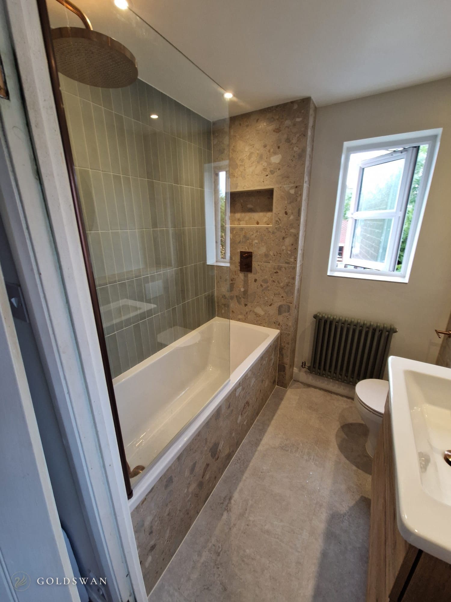 Bath with a fixed glass screen, brushed copper rain head and concealed valve, stacked sage green tiles on the back wall and a terrazzo effect end wall with a recessed shelf