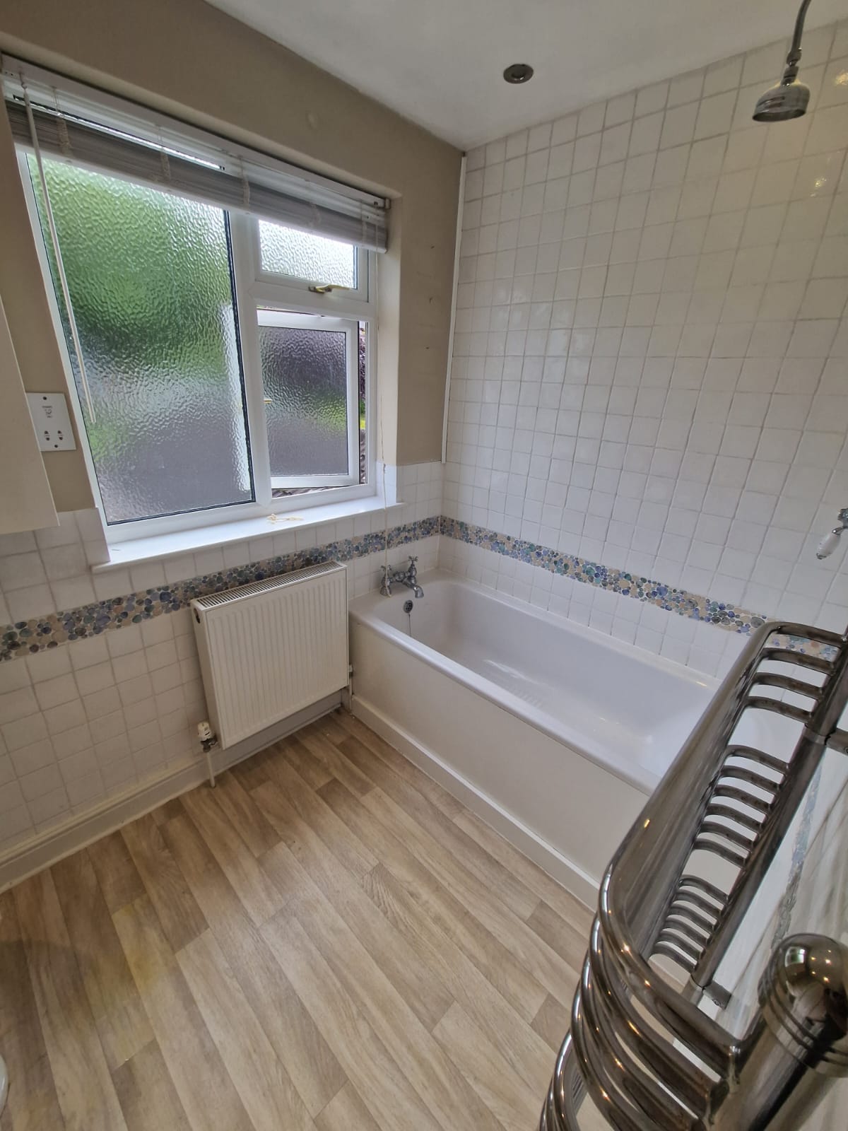 The bathroom before the work: a panel bath, white square tiles with a pebble mosaic border, and laminate flooring