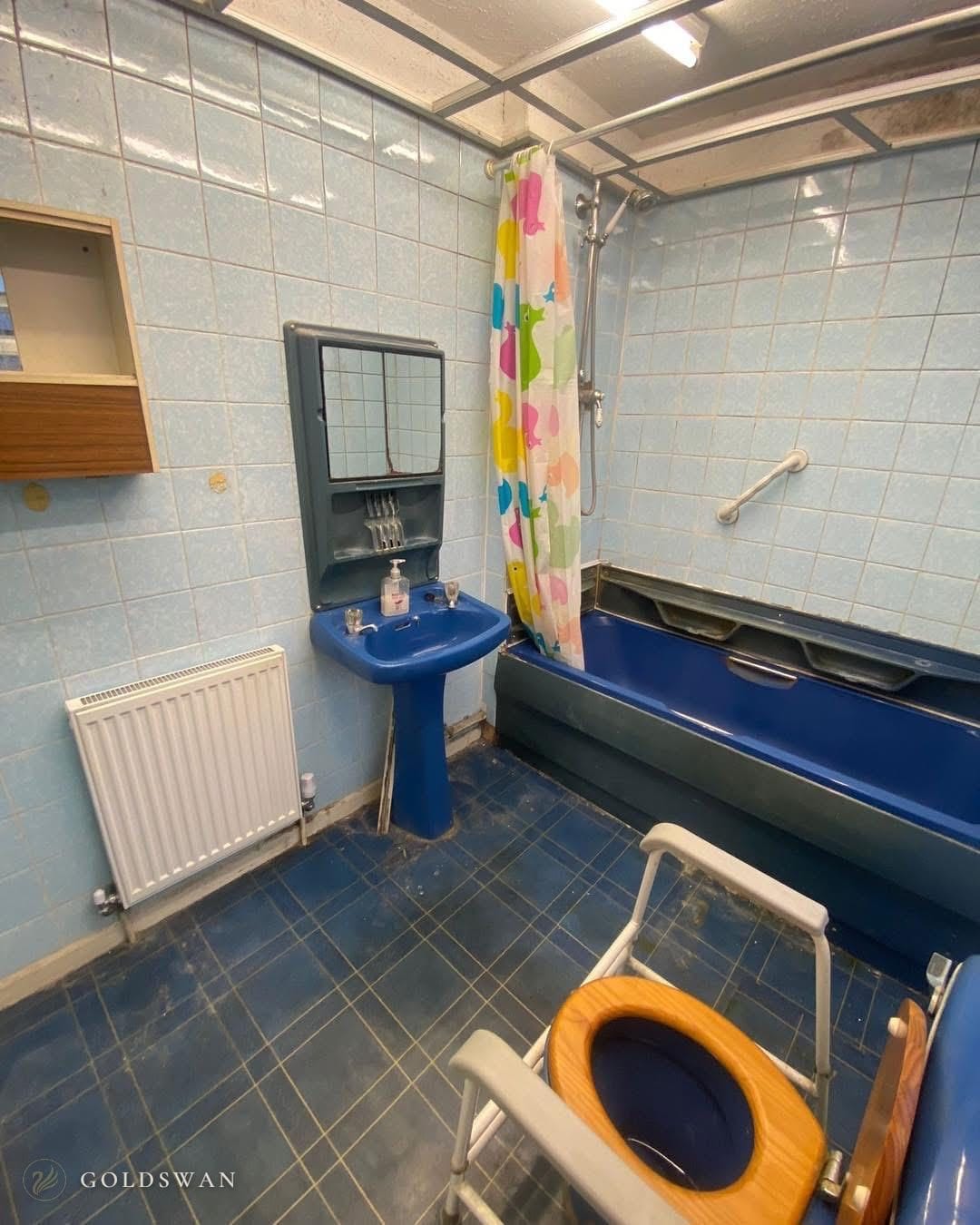 The bathroom before: a blue bath with a shower curtain, a blue pedestal basin, pale blue wall tiles and a dark blue tiled floor