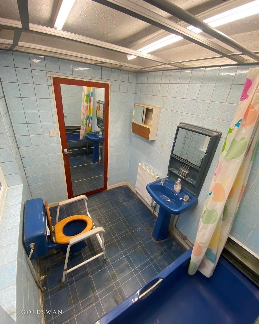 The bathroom before, looking towards the door: a blue WC with a toilet frame, a blue pedestal basin and a strip-lit grid ceiling