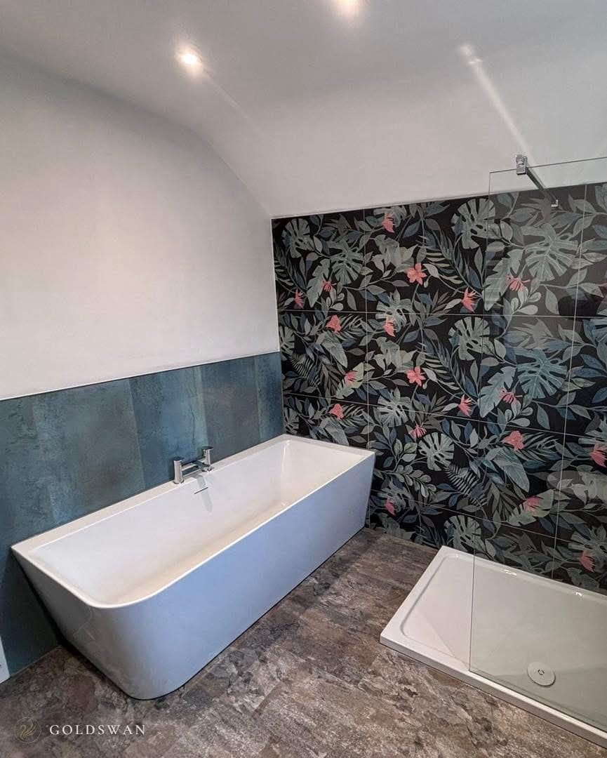 Back to wall freestanding bath with a chrome mixer against teal metallic tile, beside a black tropical leaf tile wall with pink flowers