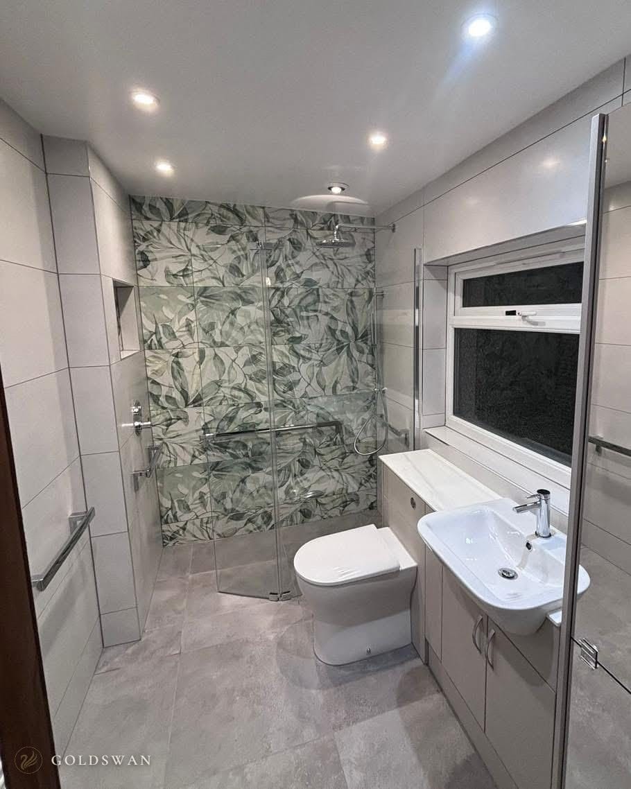 Wet room with a level grey stone effect floor, a green leaf tiled shower wall behind a hinged glass screen, chrome grab rails, a back to wall WC and a semi-recessed basin on pale fitted units