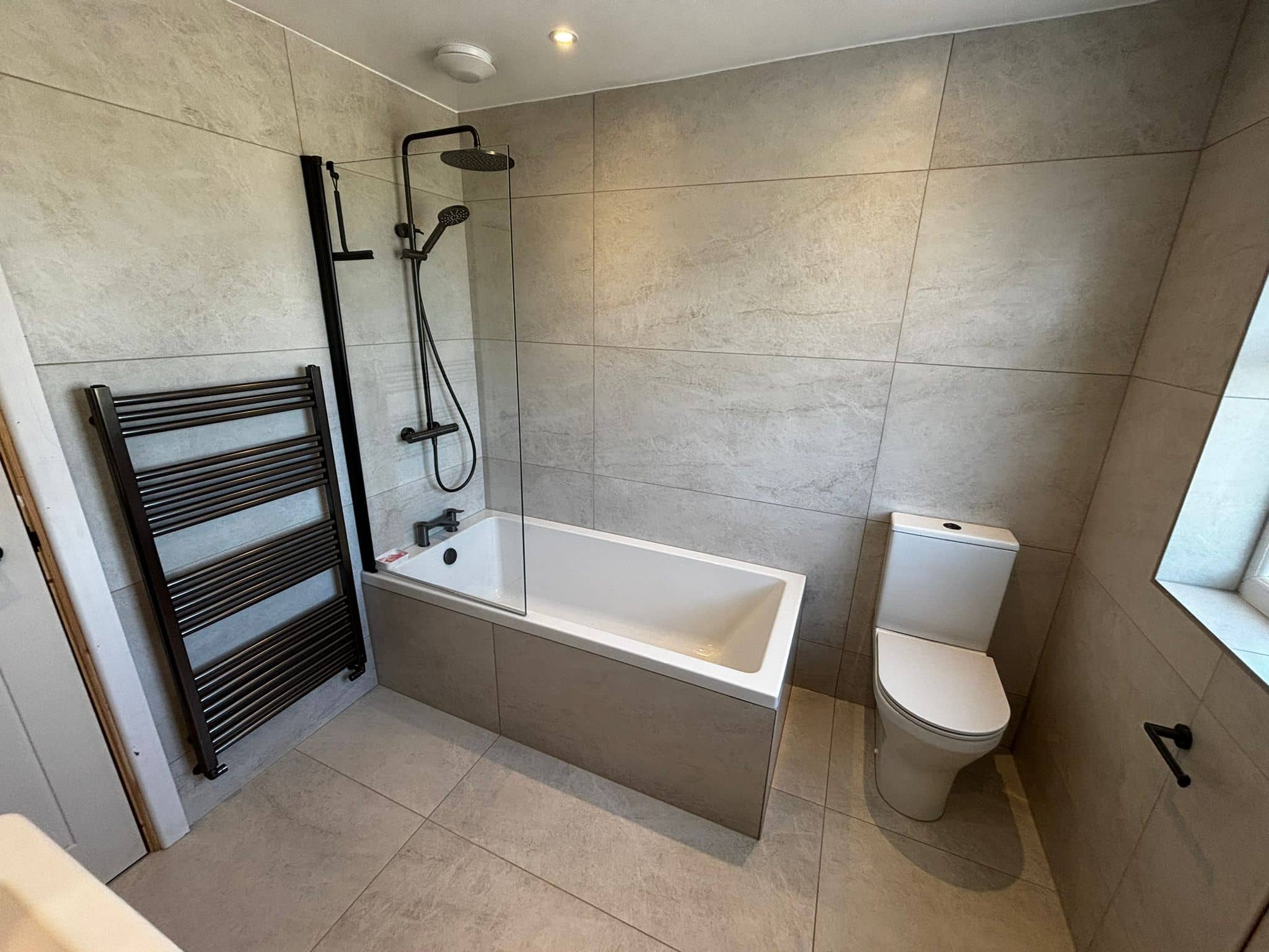 Bath with a matt black rainfall shower over it, black towel rail and stone-effect tiles