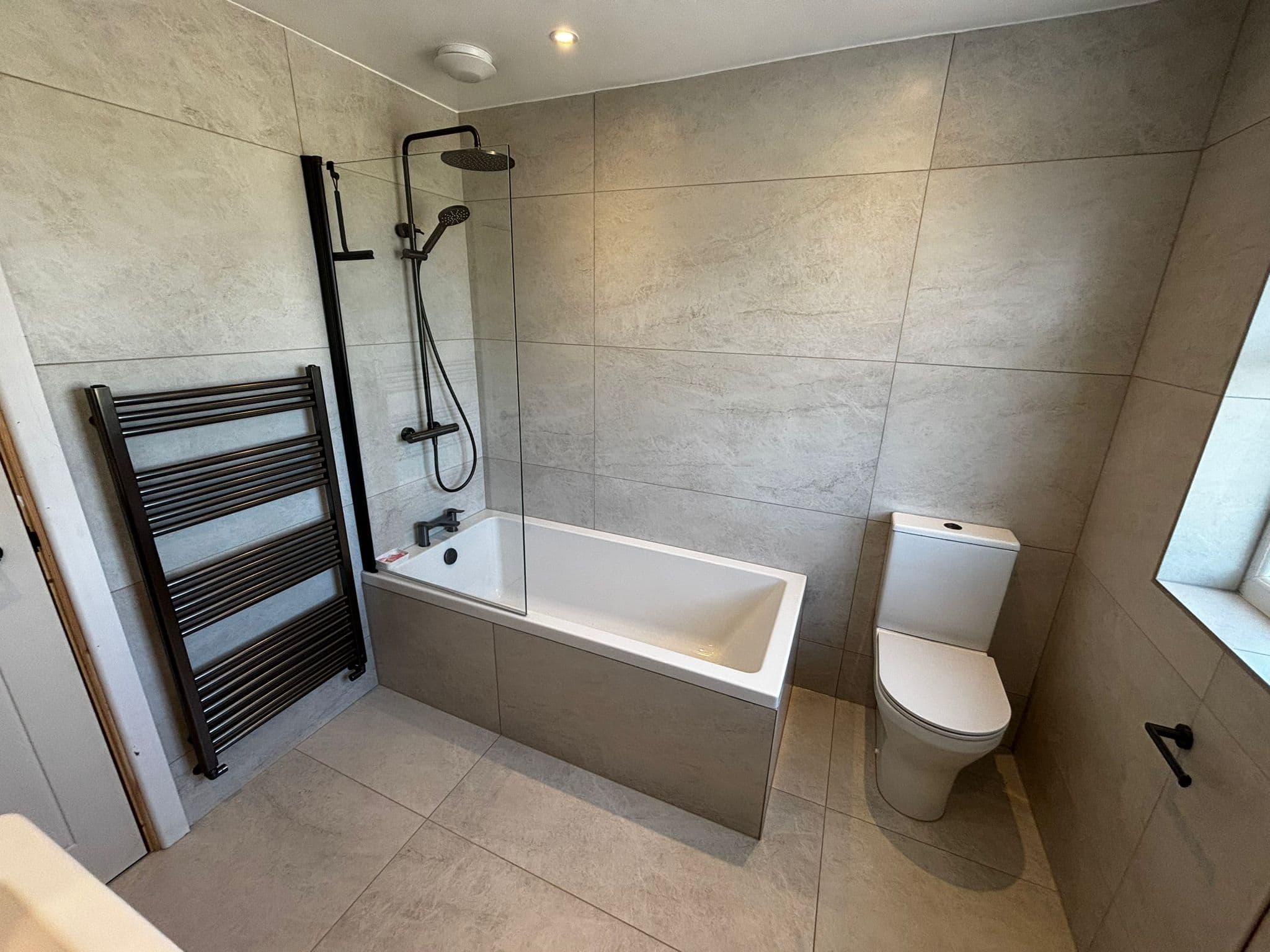Bath with a matt black rainfall shower over it, black towel rail and stone-effect tiles