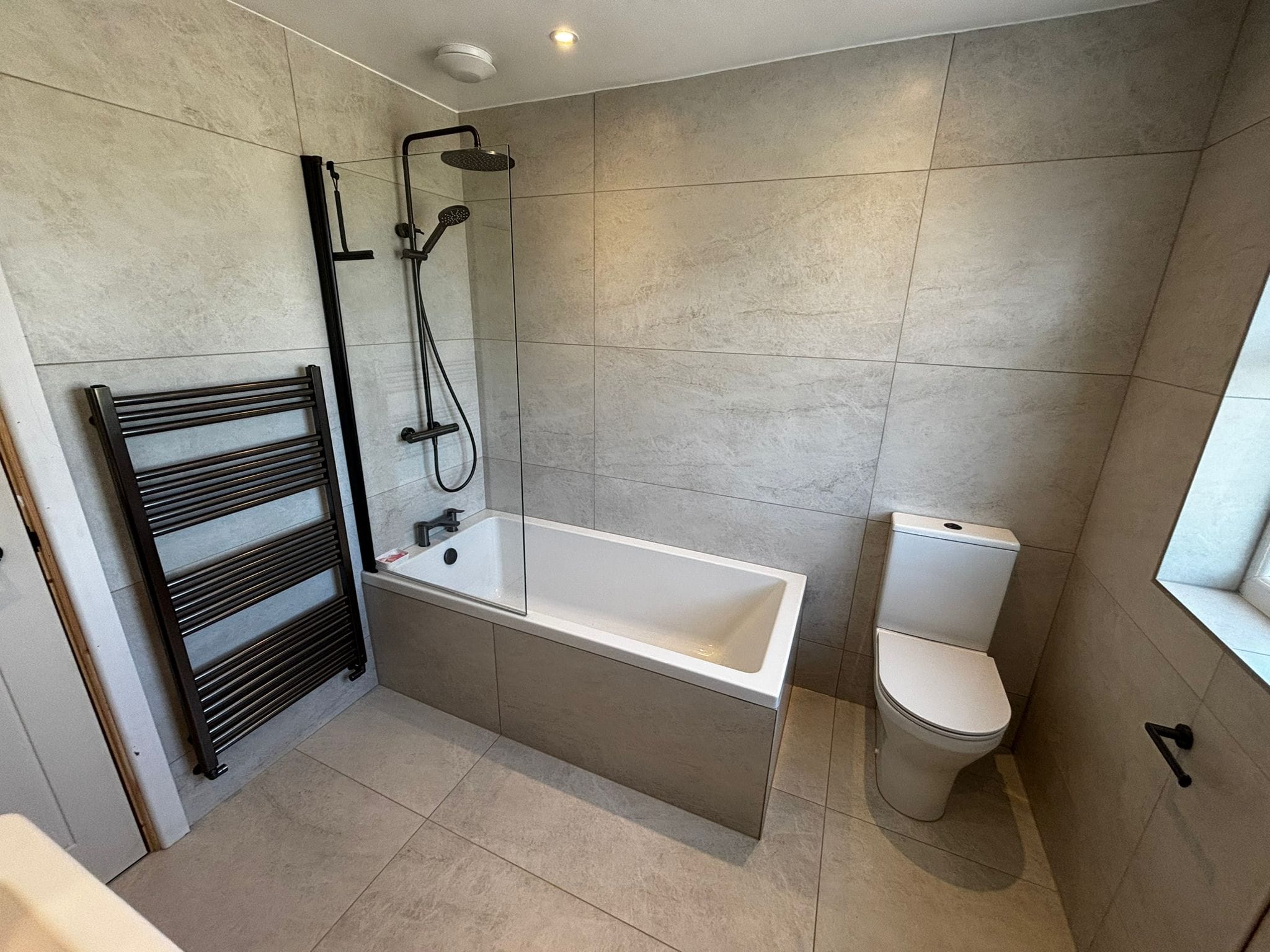 Bath with a matt black rainfall shower over it, black towel rail and stone-effect tiles