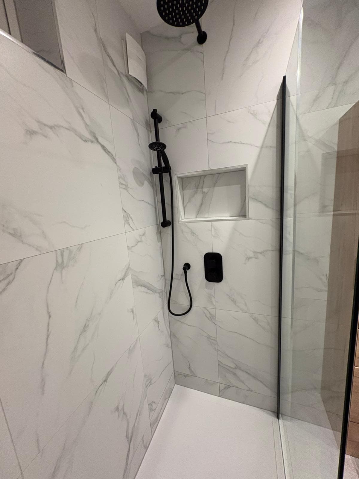 Black shower valve and slide rail beside a recessed tiled niche in marble walls