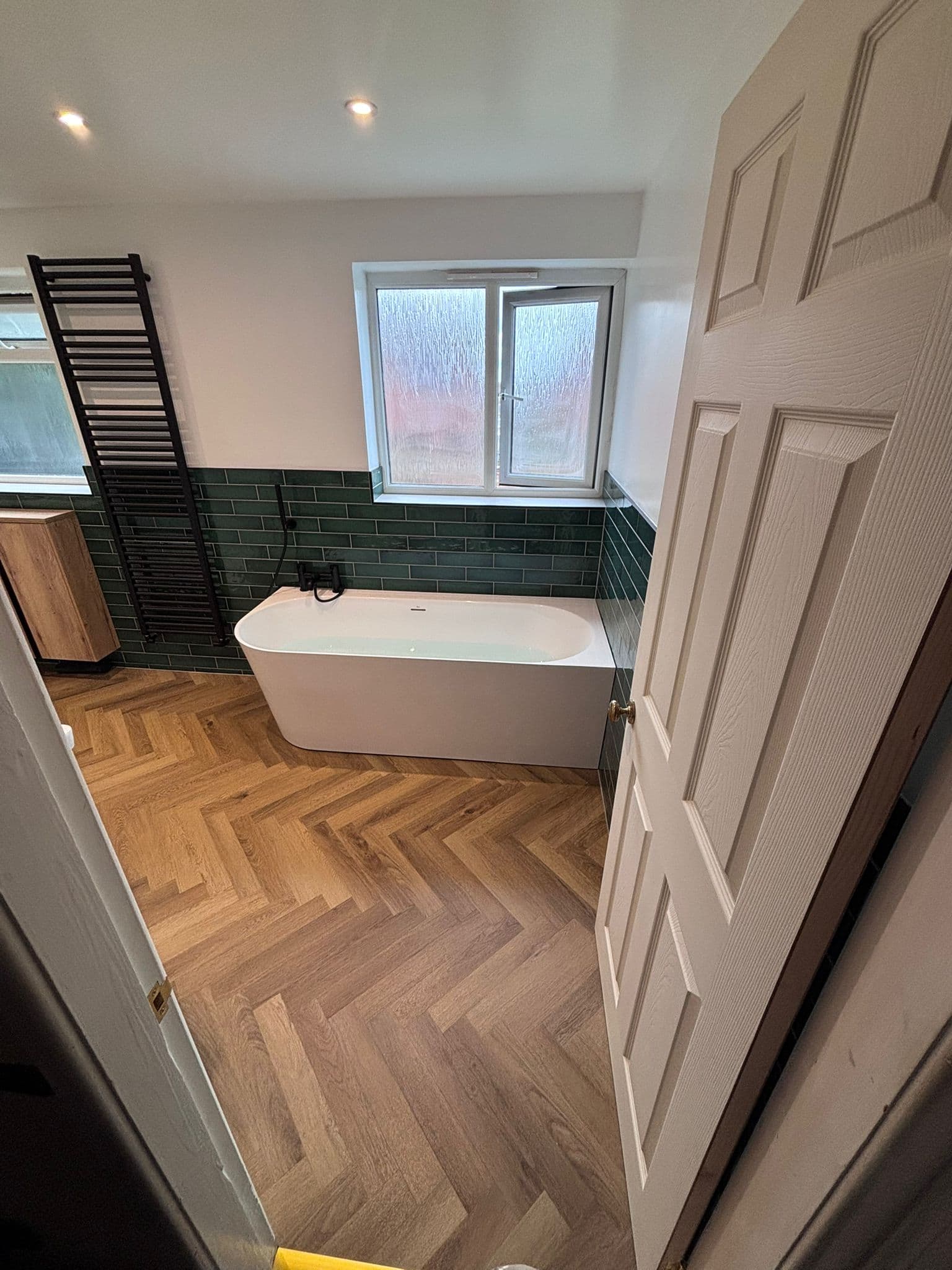 Freestanding bath under the window framed by green tiles and a black towel rail