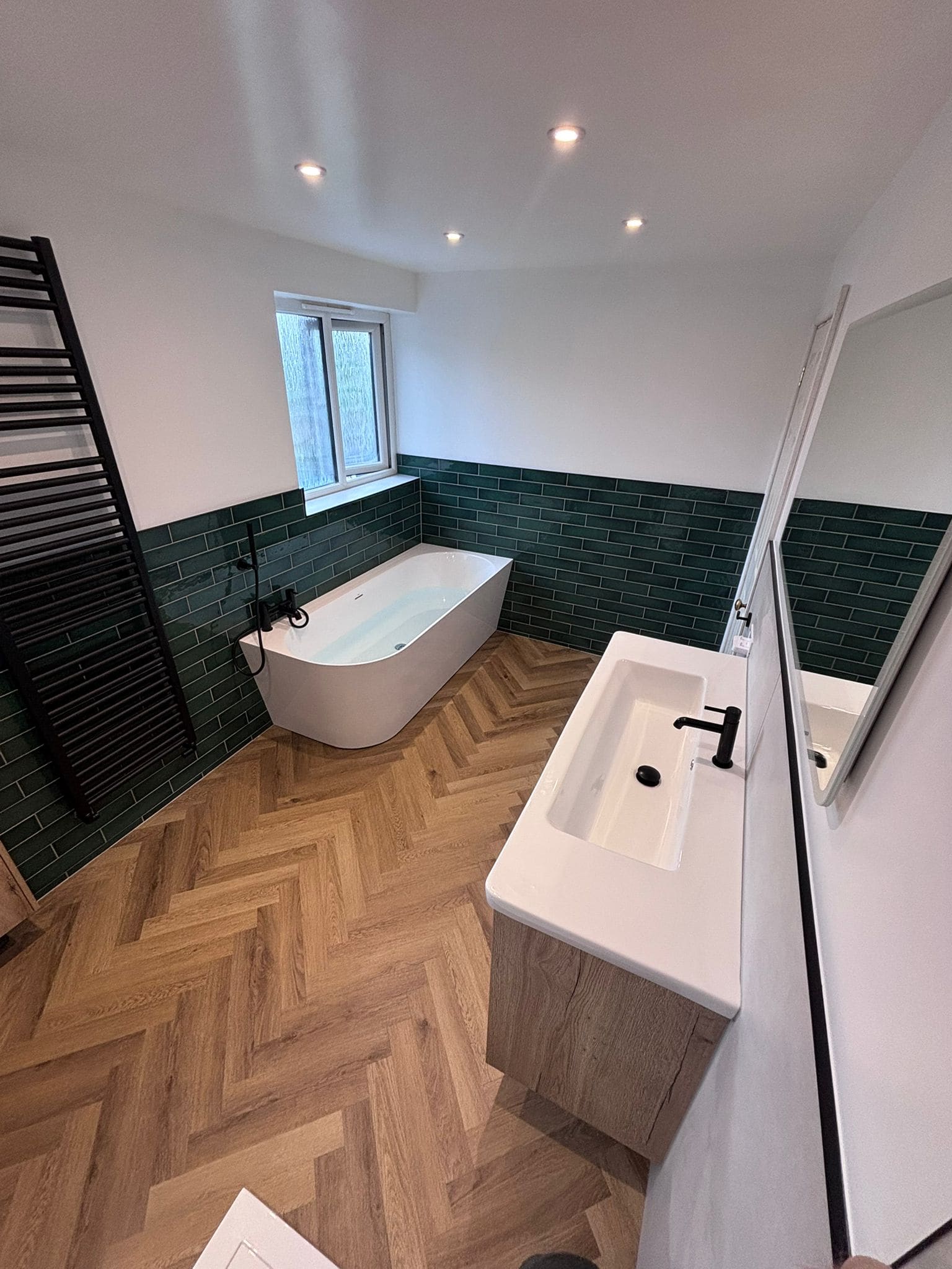 Freestanding bath against green tiles with an oak vanity in the foreground