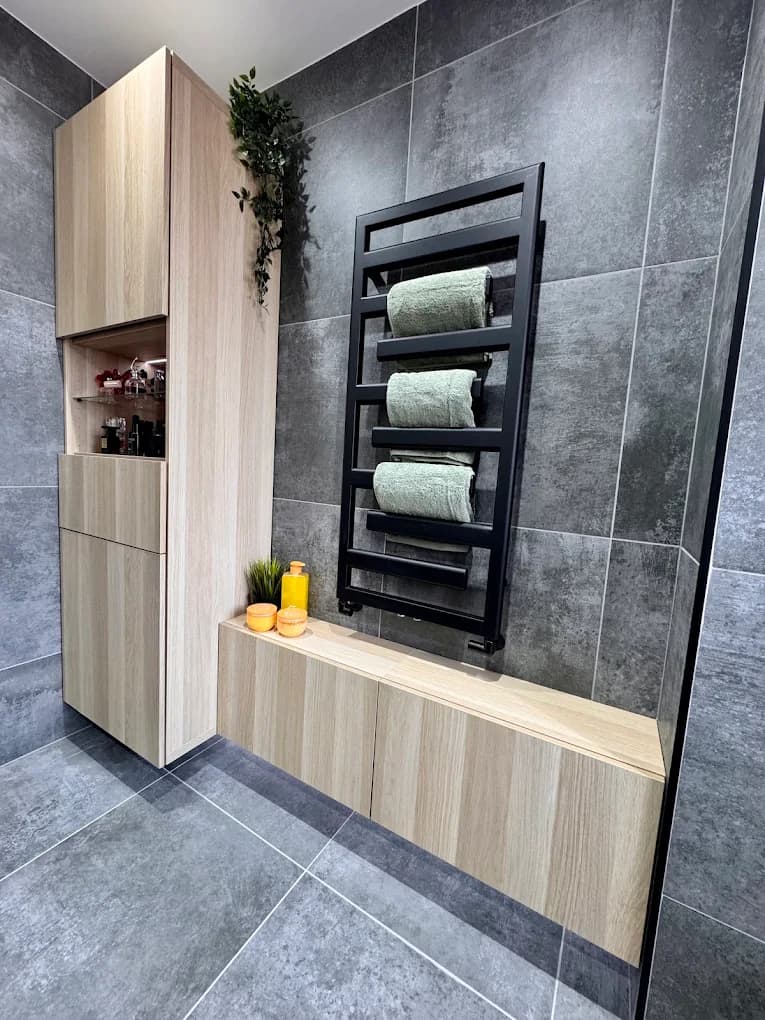 Black towel radiator holding green towels against grey tiles and a light oak storage bench