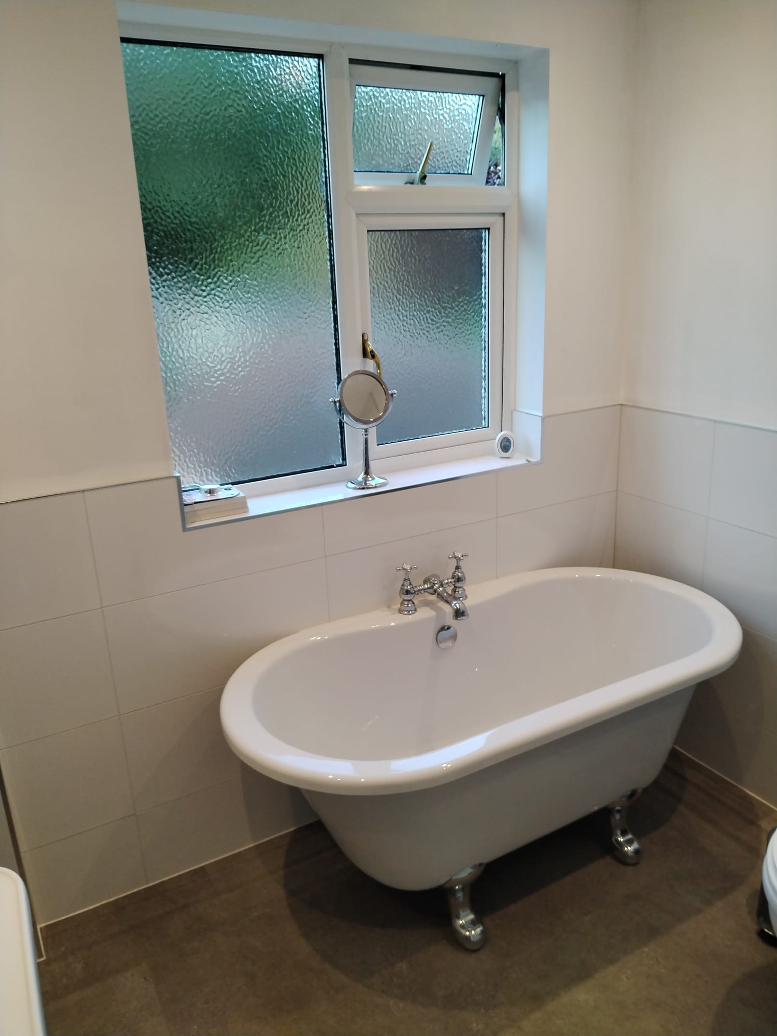 Freestanding roll top bath on chrome feet, under an obscured glass window, with cream wall tiles and a dark stone effect floor