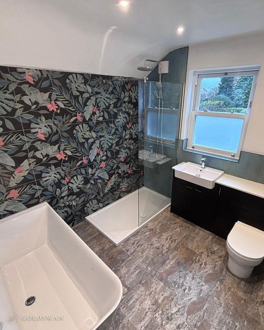 Tropical leaf tile wall behind a walk-in shower with a fixed glass panel, a back to wall freestanding bath, dark fitted furniture with a semi-recessed basin and a slate effect floor