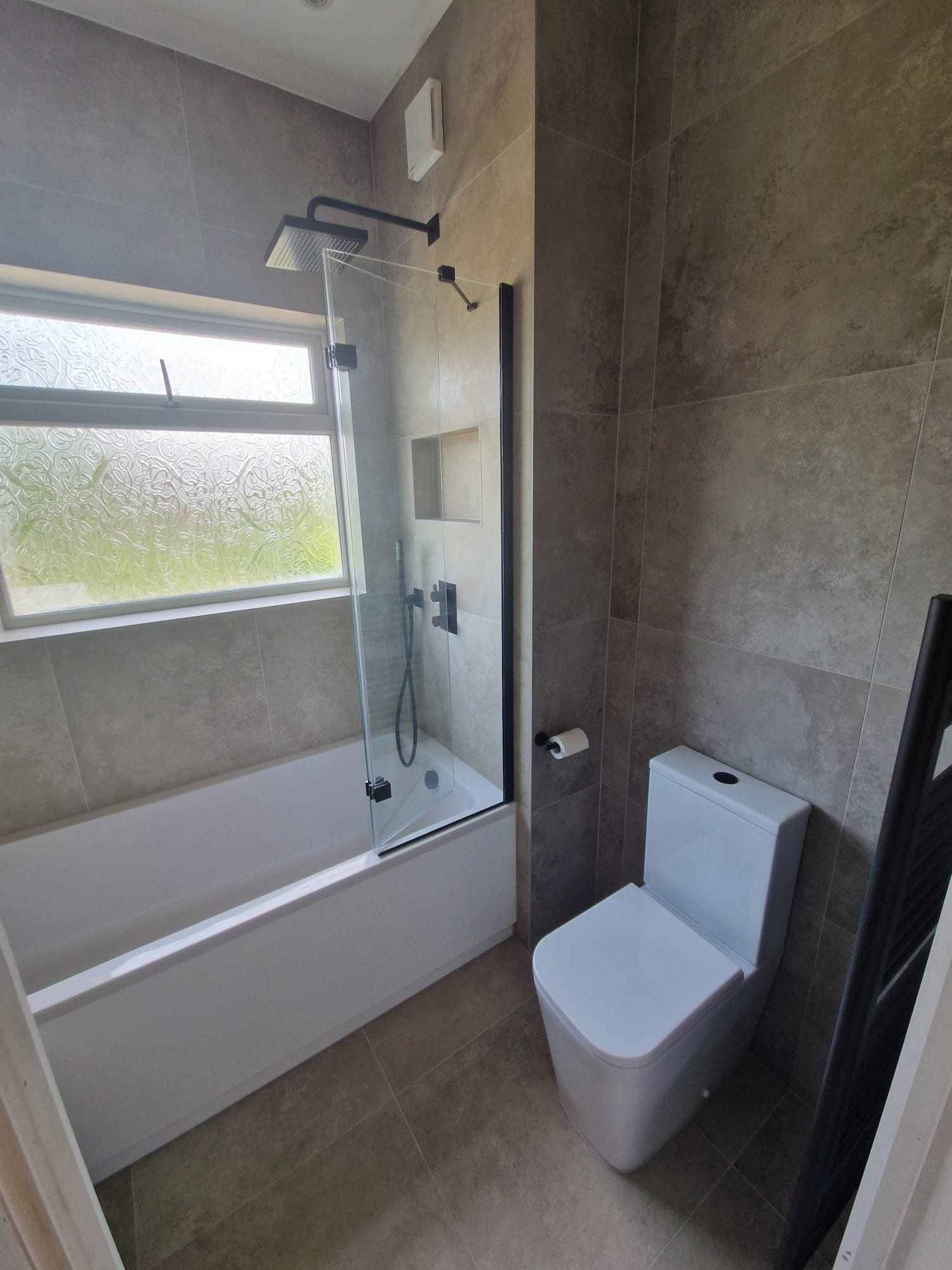 Bath with a black framed shower screen and a black rain head, against grey stone effect tiles with a recessed shelf