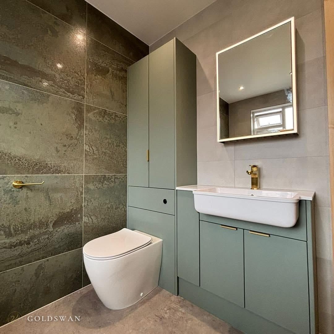 Sage green fitted furniture with a tall cupboard, a unit hiding the cistern of a back to wall toilet, and a vanity with a semi-recessed basin and a brushed brass tap under an illuminated square mirror, beside a dark green oxidised effect tiled wall