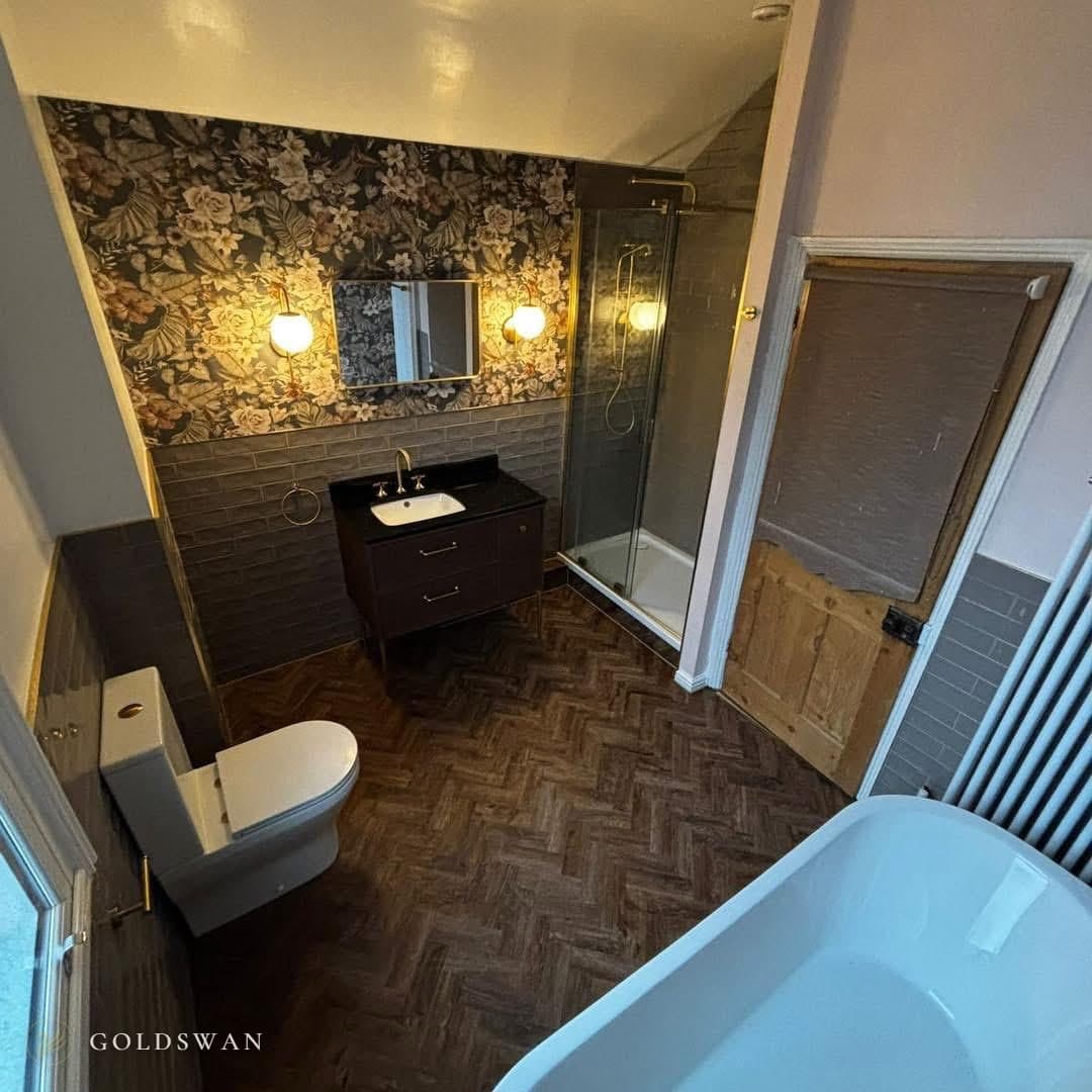 The same bathroom after: a dark vanity on legs with drawers and a brass tap under a brass framed mirror between two globe wall lights, a dark floral wall above grey metro tile, a glass shower enclosure tiled to the ceiling beside the same wooden door, a herringbone wood effect floor and the end of a freestanding bath
