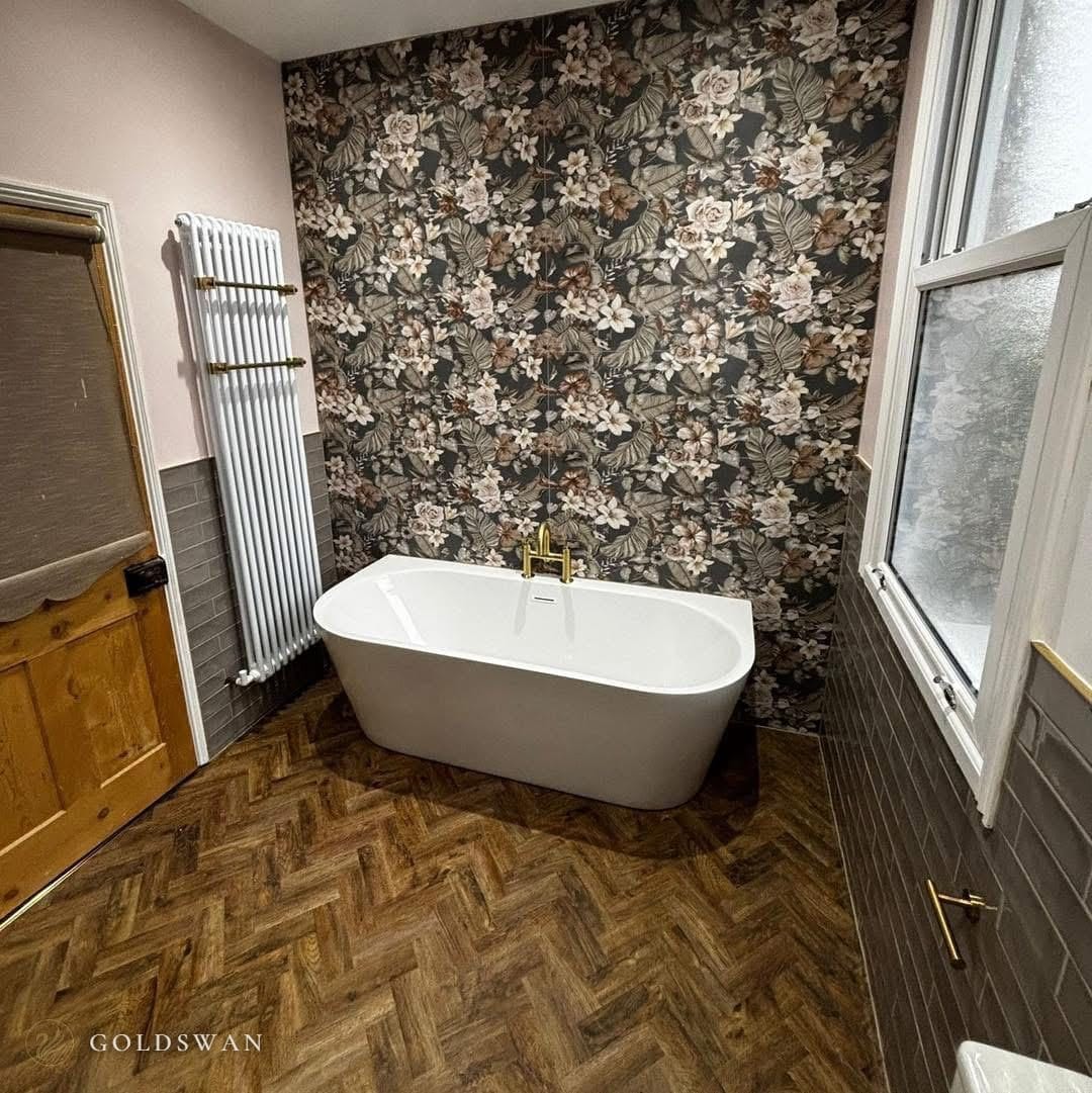 Back to wall freestanding bath with a brushed brass mixer against a floor to ceiling dark floral tile, with a herringbone wood effect floor and a white column radiator