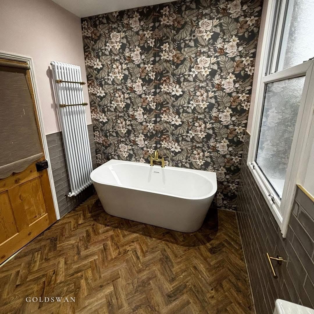 Back to wall freestanding bath with a brushed brass mixer against a floor to ceiling dark floral tile, with a herringbone wood effect floor and a white column radiator