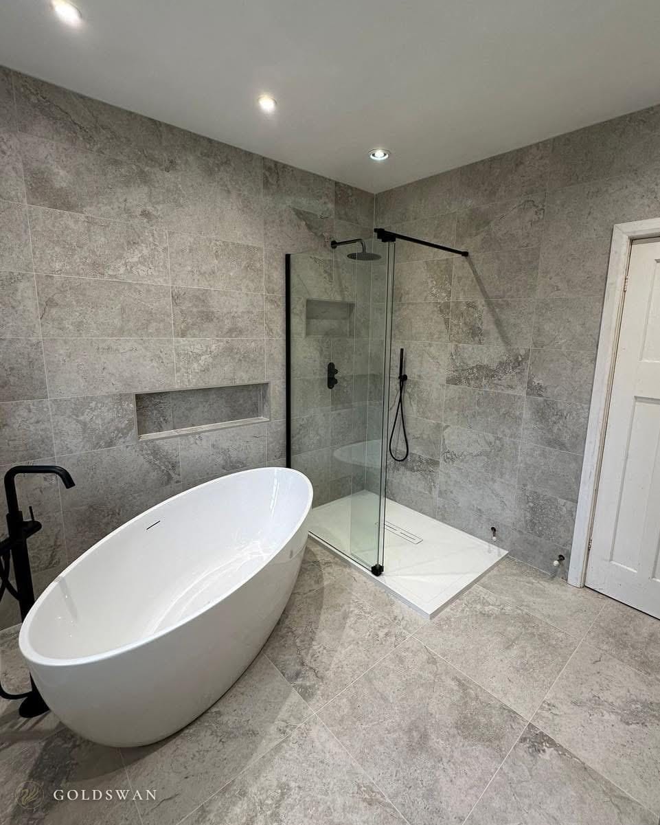 Oval freestanding bath with a matt black floor standing filler, beside a walk-in shower with a black framed glass panel, in grey stone effect tile with a long recessed shelf