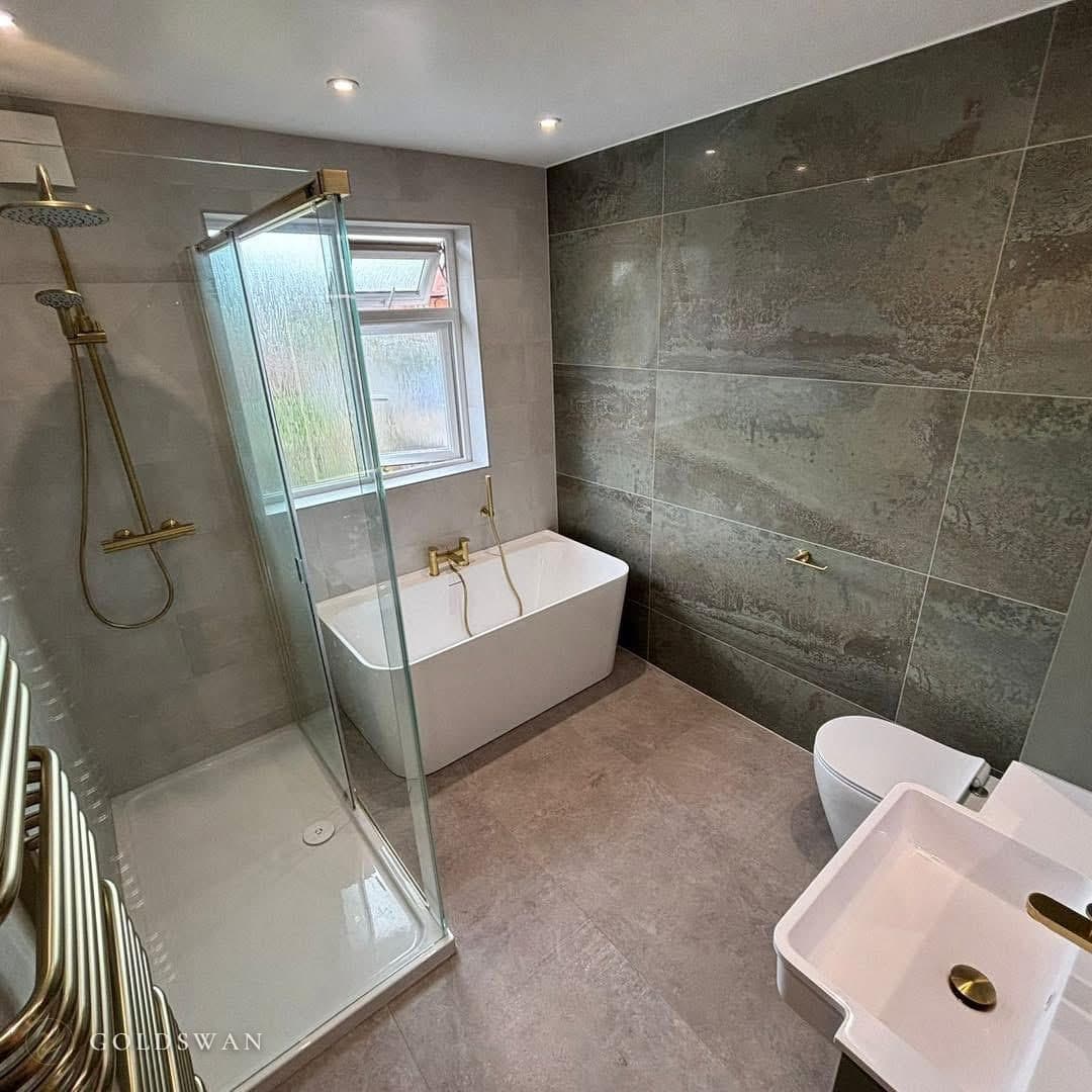 Back to wall freestanding bath under a window with a brushed brass deck mixer and handset, beside a glass shower enclosure with a low white tray and a brushed brass riser, with a dark green oxidised effect feature wall and a brushed brass towel radiator
