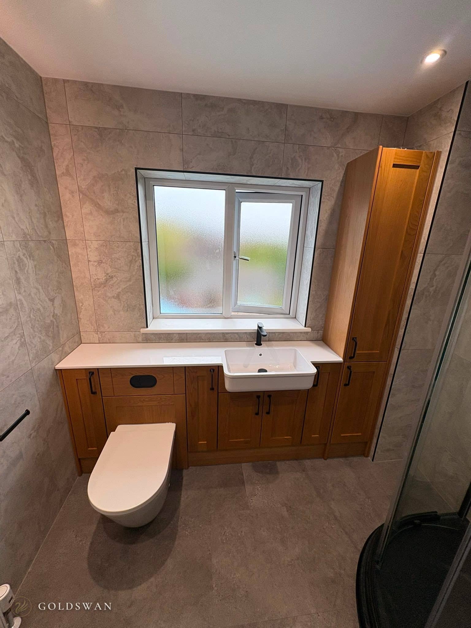 Solid oak shaker furniture under a frosted window, with a semi-recessed basin, matt black tap, a back to wall WC with a black flush plate and a tall oak cupboard