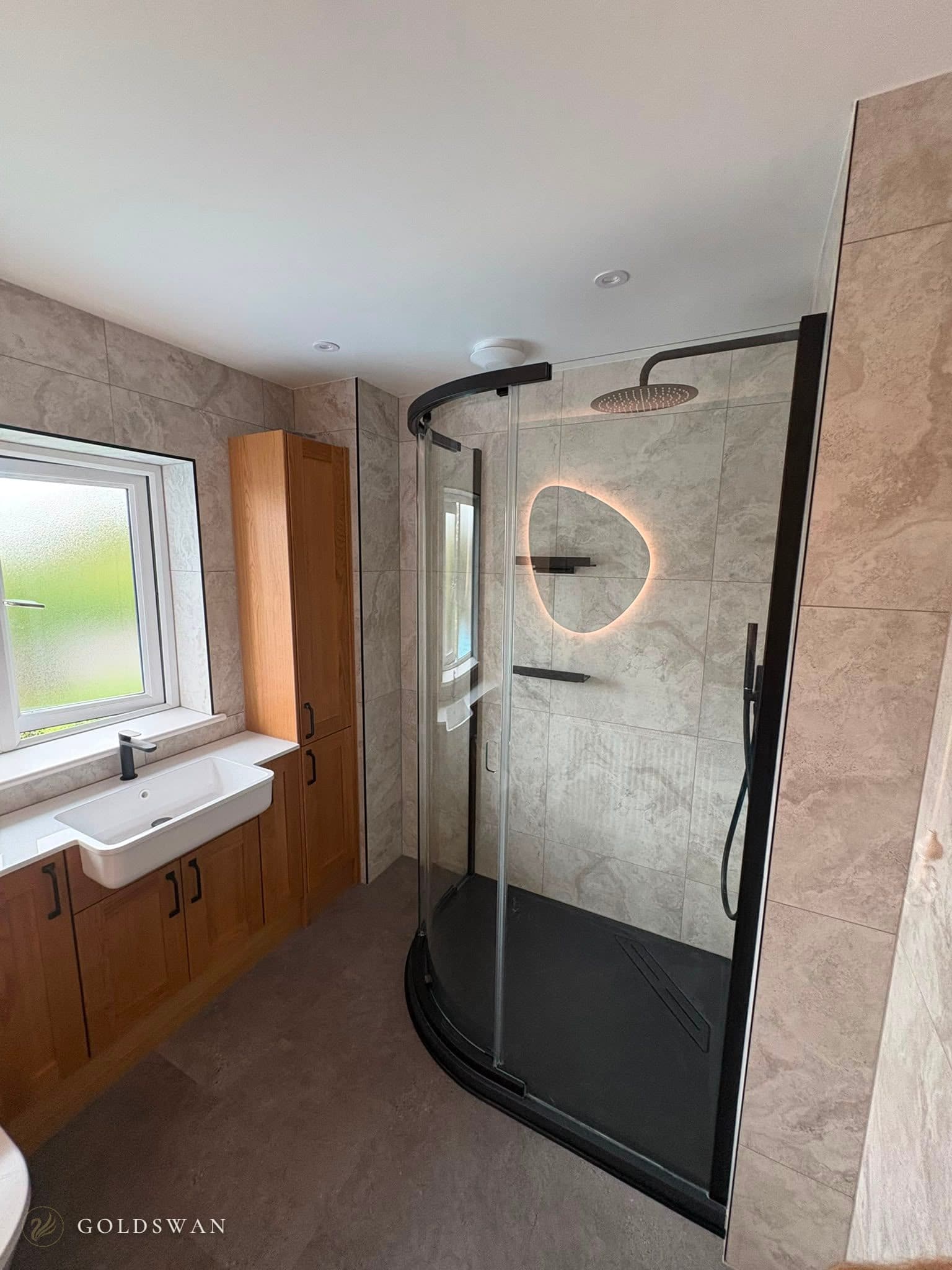 Curved black framed shower enclosure with a black slate effect tray, matt black rain head and handset, beside a solid oak vanity run with a semi-recessed white basin, all in grey stone effect tile